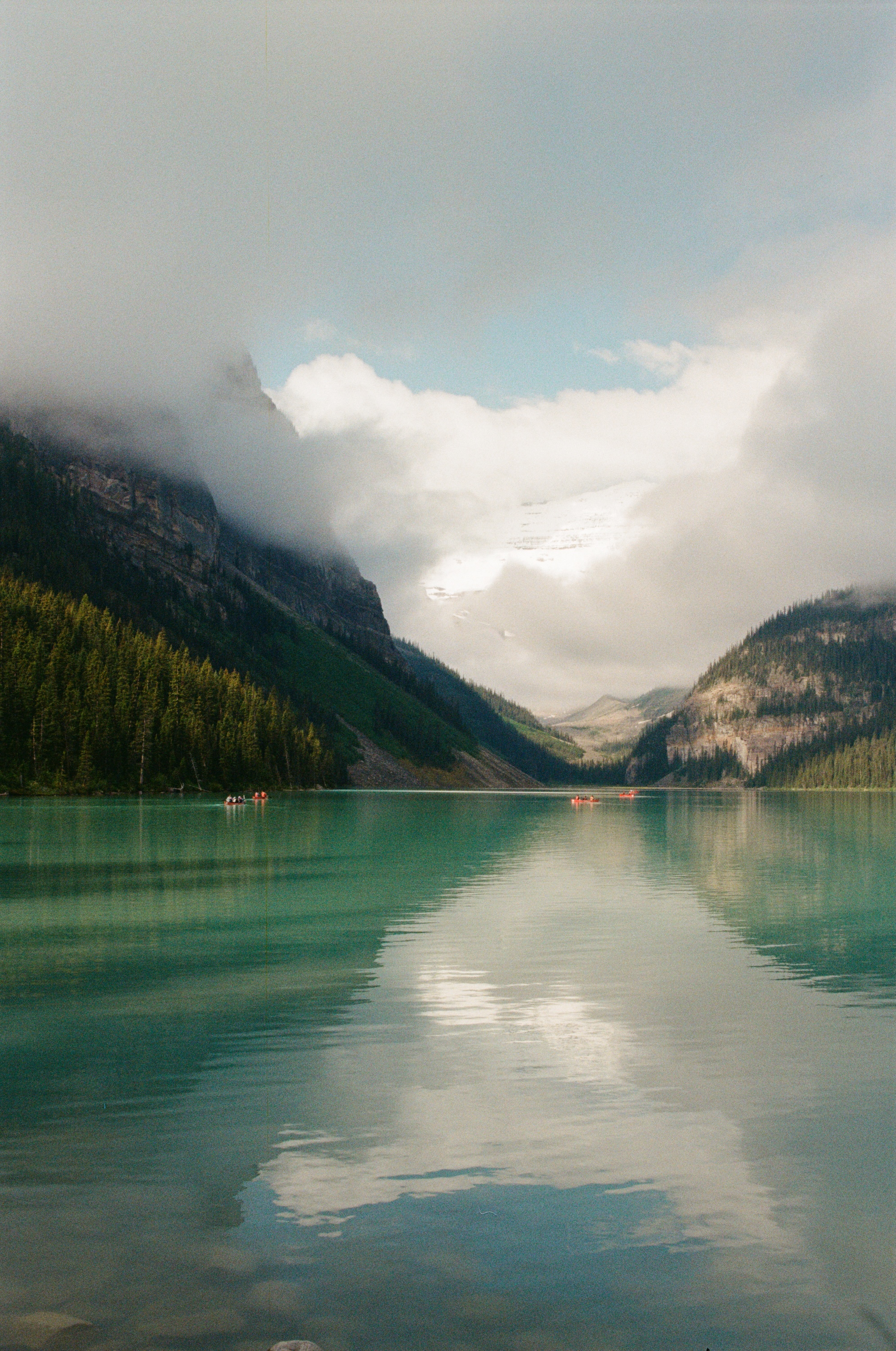 Alpine lake at sunrise, clouds spilling down a mountain pass, two red canoes drifting on the water.